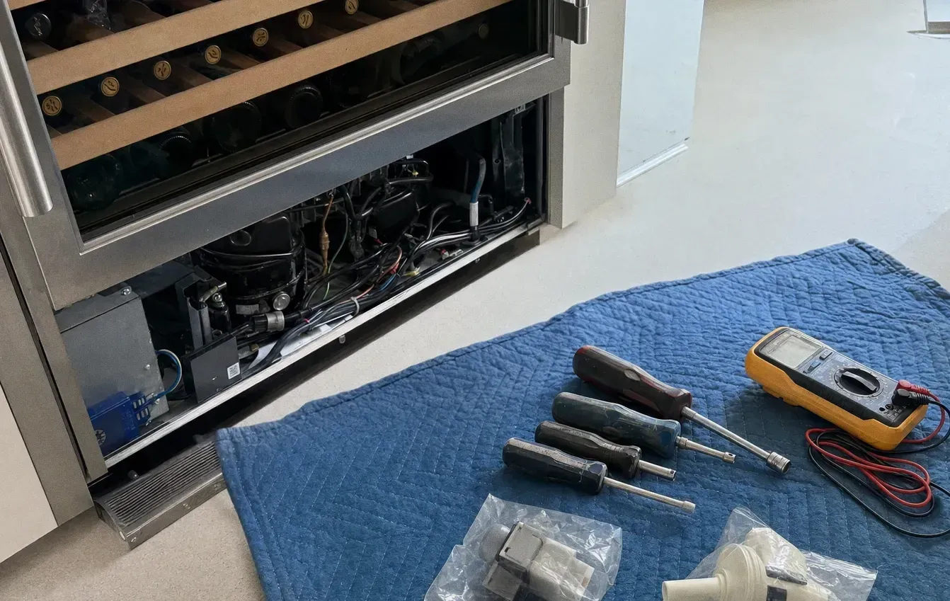Stainless wine cabinet with wooden racks above and its lower machine compartment open, screwdrivers, a multimeter and small parts laid out on a mat